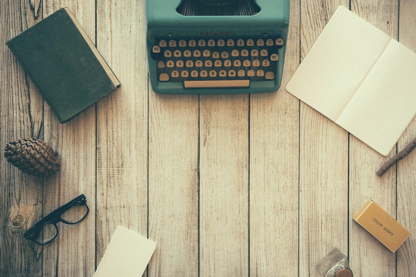 A teal vintage typewriter on a wooden table, surrounded by a closed book, an open notebook, glasses, and a pine cone.