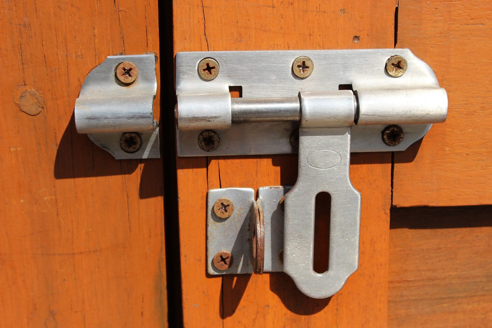 A metal barrel bolt latch on an orange wooden door, slid open and unlocked.