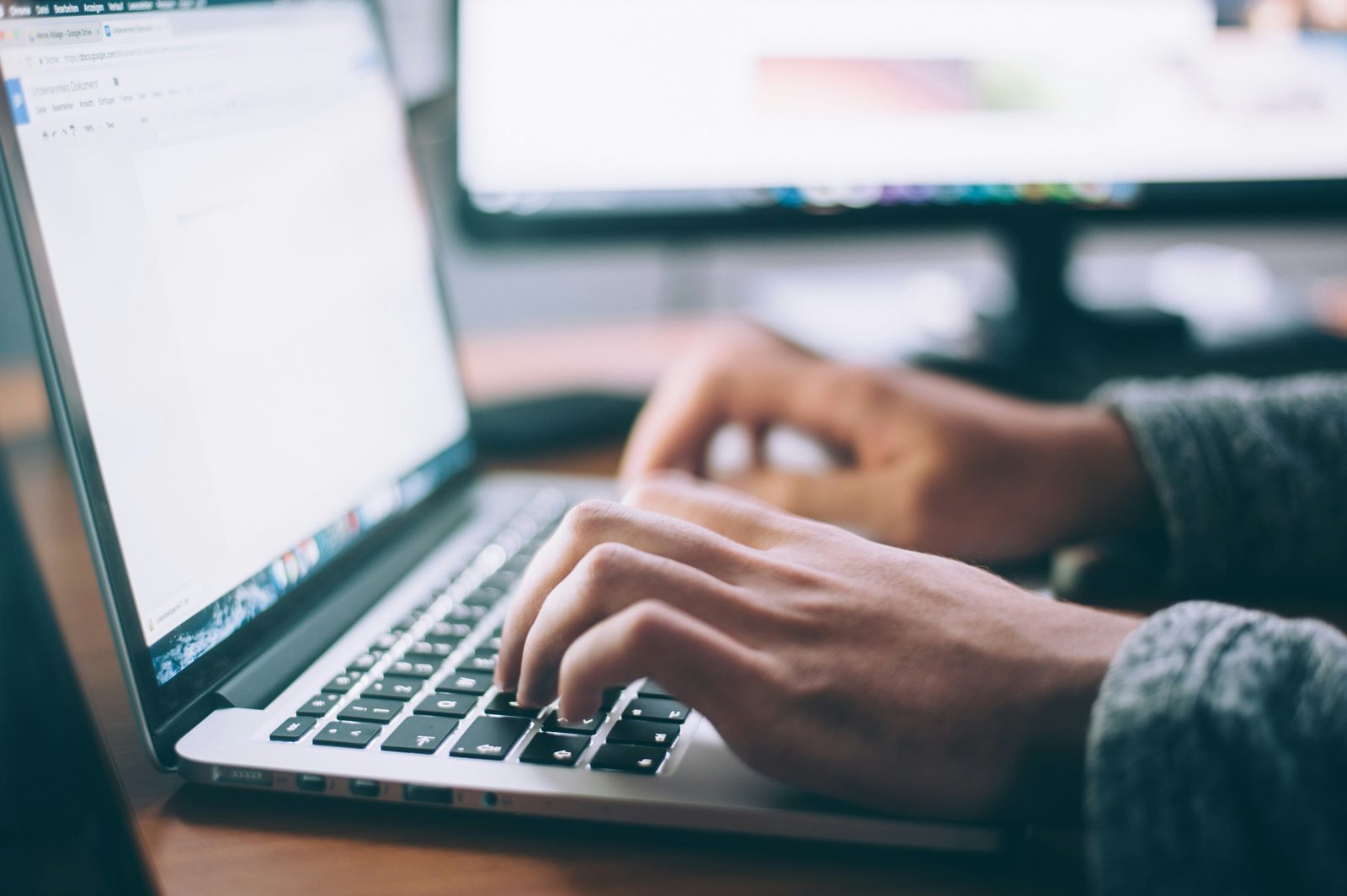 Close-up of hands typing on a laptop keyboard, with a document open on the screen and a monitor in the background.