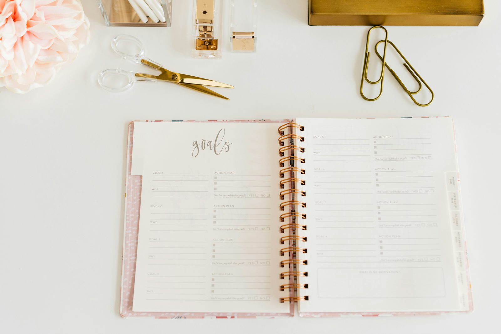 An open spiral planner on a white desk, with a page titled goals divided into goal and action plan sections, next to gold scissors, a stapler, and paper clips.