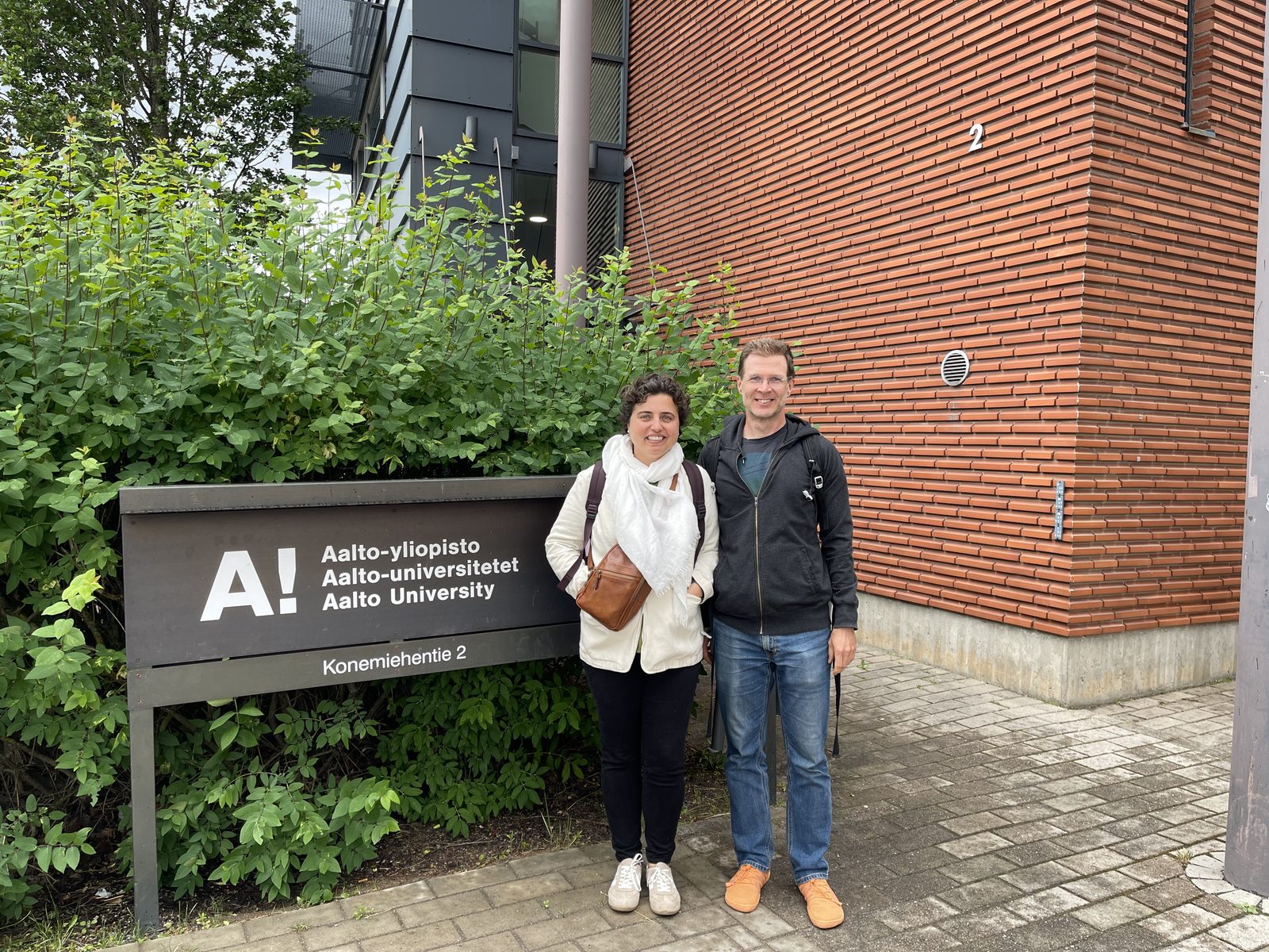 A man and a woman standing next to a sign that reads Aalto University, with the university's name written first in Finnish, then in Swedish, and then in English.