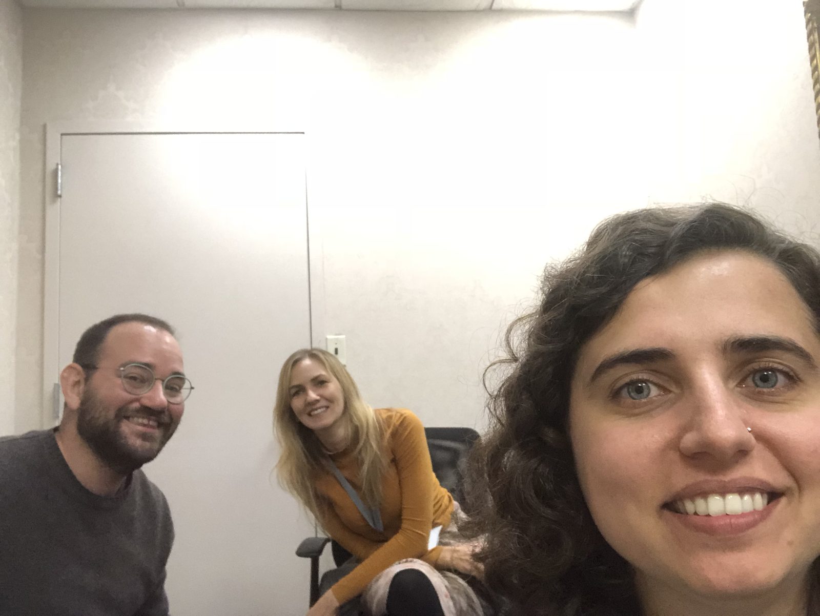 Selfie of three people smiling. From left to right: a man with glasses, a beard, and a grey sweater; a woman with blonde hair and a mustard-colored sweater; and, on the right, a woman with curly hair taking the selfie.