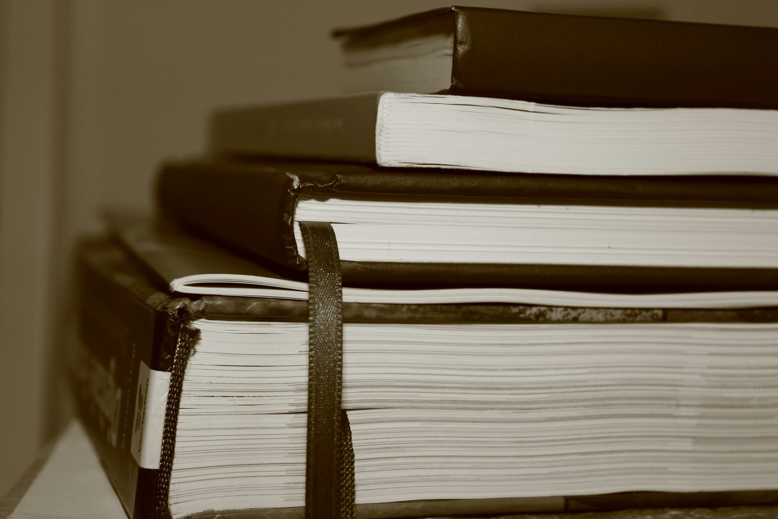 A sepia-toned close-up of a stack of thick hardcover books, one with a ribbon bookmark.